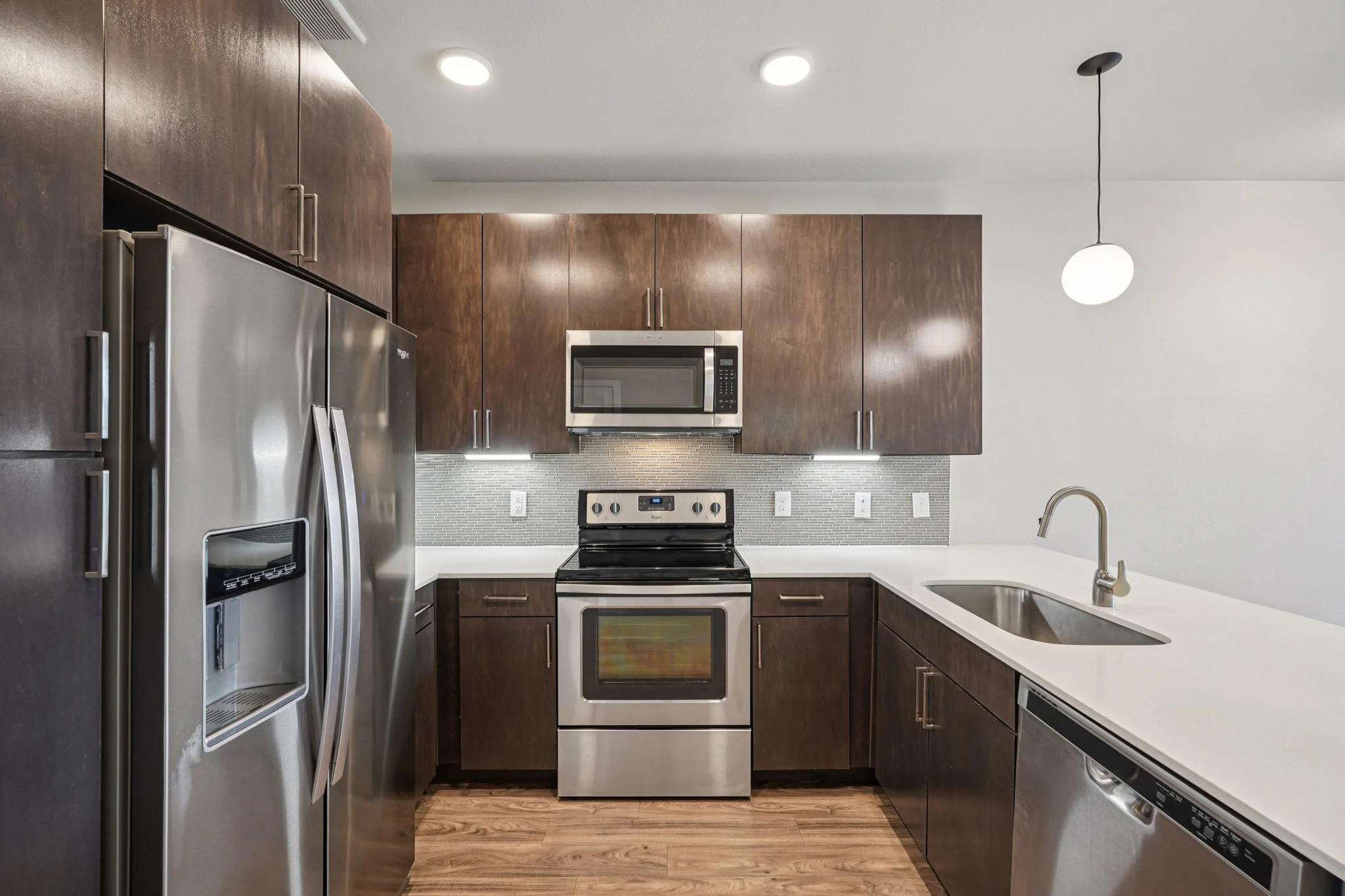 A modern kitchen featuring stainless steel appliances, dark wood cabinets, and a white countertop. The layout includes a refrigerator, microwave, oven, and a sink with a sleek faucet.