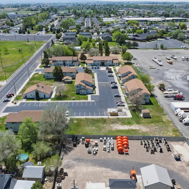 Aerial View of Community and Industrial Area An aerial view of a residential community alongside an industrial area. The image includes several houses, parking lots, and commercial vehicles in a nearby lot, with a recreational park in the background.
