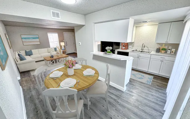Contemporary Dining and Kitchen Area A bright and modern dining area connected to a kitchen and seating area. A round wooden table is set with white plates and a pink flower centerpiece. The kitchen features white cabinets and a window with a view of the adjacent room. The living room can be seen in the background with a white sofa and coffee table.