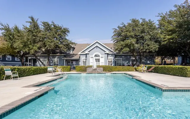 A clear swimming pool surrounded by lounge chairs and lush greenery, with residential buildings in the background.