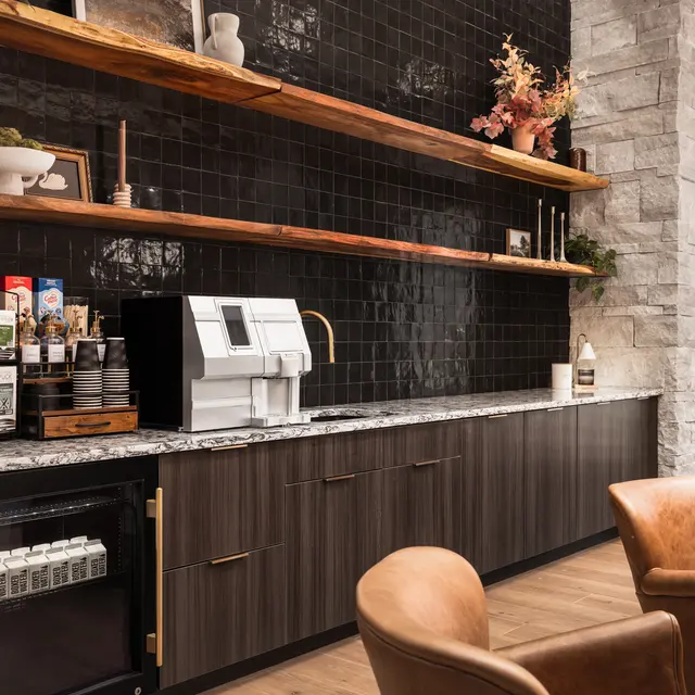 A modern kitchenette featuring a dark tiled wall, wooden shelves filled with decorative items, a coffee machine, and various coffee supplies. Leather chairs are placed in front.