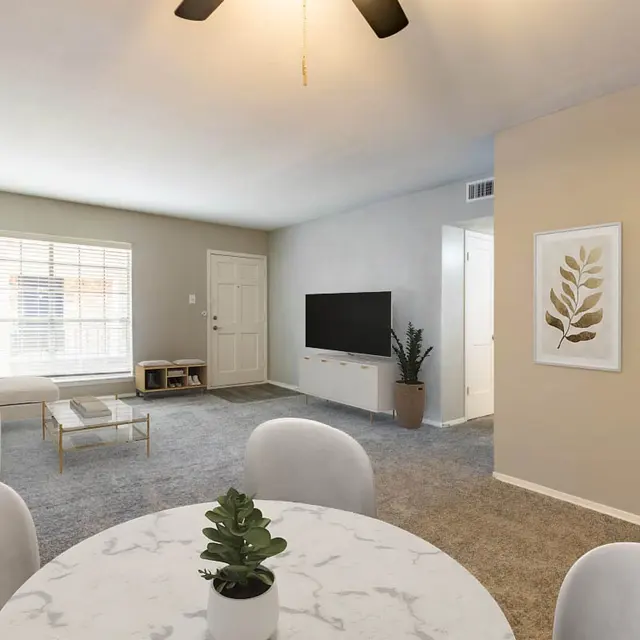 A bright and spacious living room featuring a comfortable sofa, a coffee table, and a television stand against a wall. There's natural light coming through a window, and a dining table with chairs in the foreground. Decorative plants and shelves add charm to the space.
