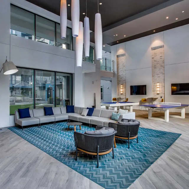 A modern lounge area featuring a sectional sofa with blue and white cushions, a round coffee table, and decorative rug. Large windows allow natural light to flow in, with a view of an outdoor area. The ceiling has hanging light fixtures, and there are two televisions mounted on the walls.