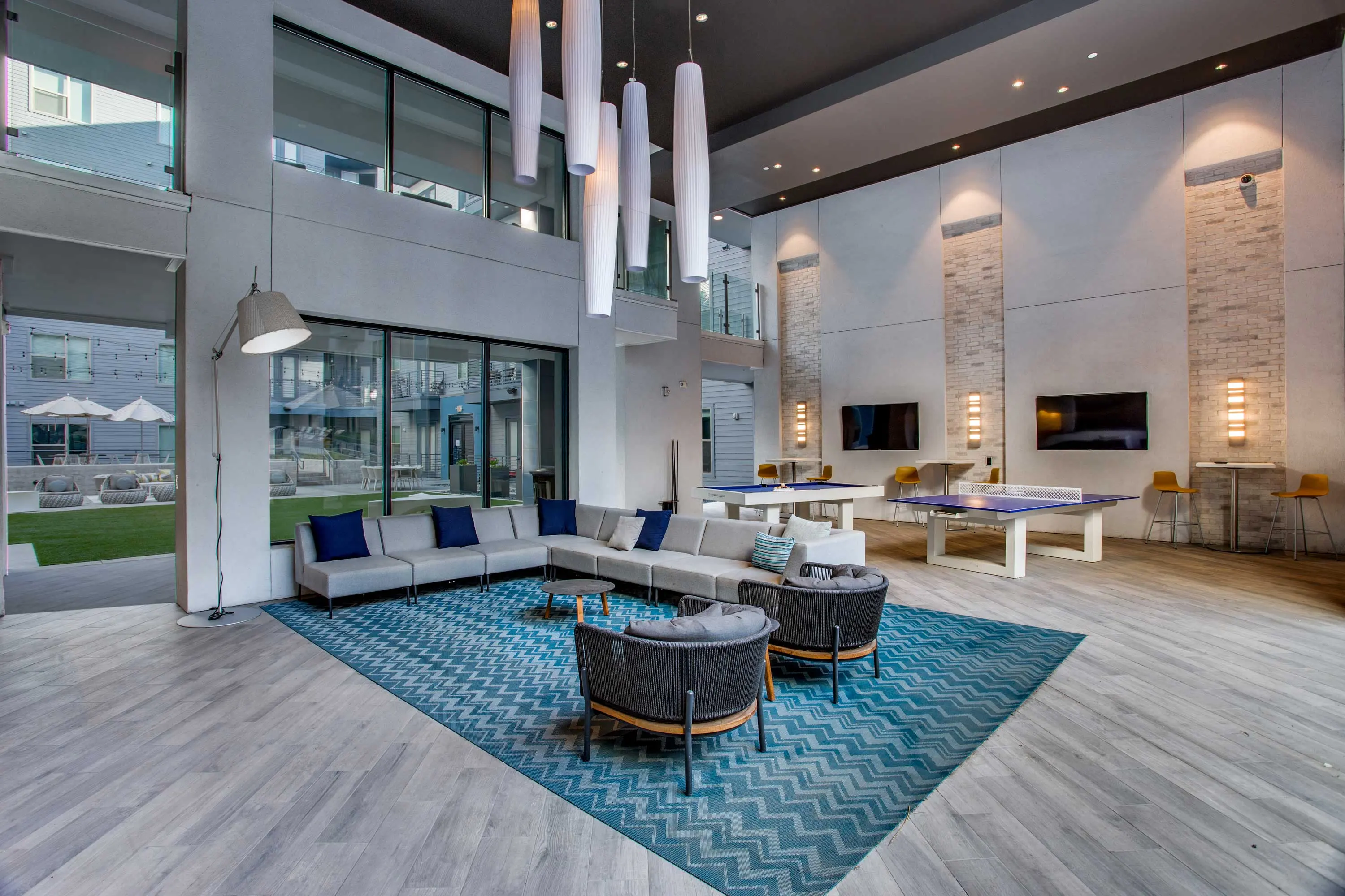 A modern lounge area featuring a sectional sofa with blue and white cushions, a round coffee table, and decorative rug. Large windows allow natural light to flow in, with a view of an outdoor area. The ceiling has hanging light fixtures, and there are two televisions mounted on the walls.