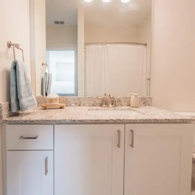 A modern bathroom featuring a granite countertop with a sink, white cabinetry, and a mirror above. The space has a light fixture with three bulbs and a towel hanging beside the sink. A shower curtain is visible in the background.