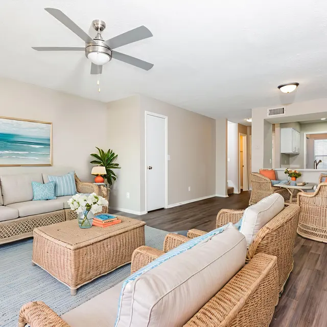 A bright and airy living room with light-colored walls and wooden flooring. It features comfortable seating with wicker sofas adorned with light blue cushions. A coffee table sits in the center, and a small plant decorates the space. A ceiling fan hangs above, and there is an open view into a kitchen area in the background.