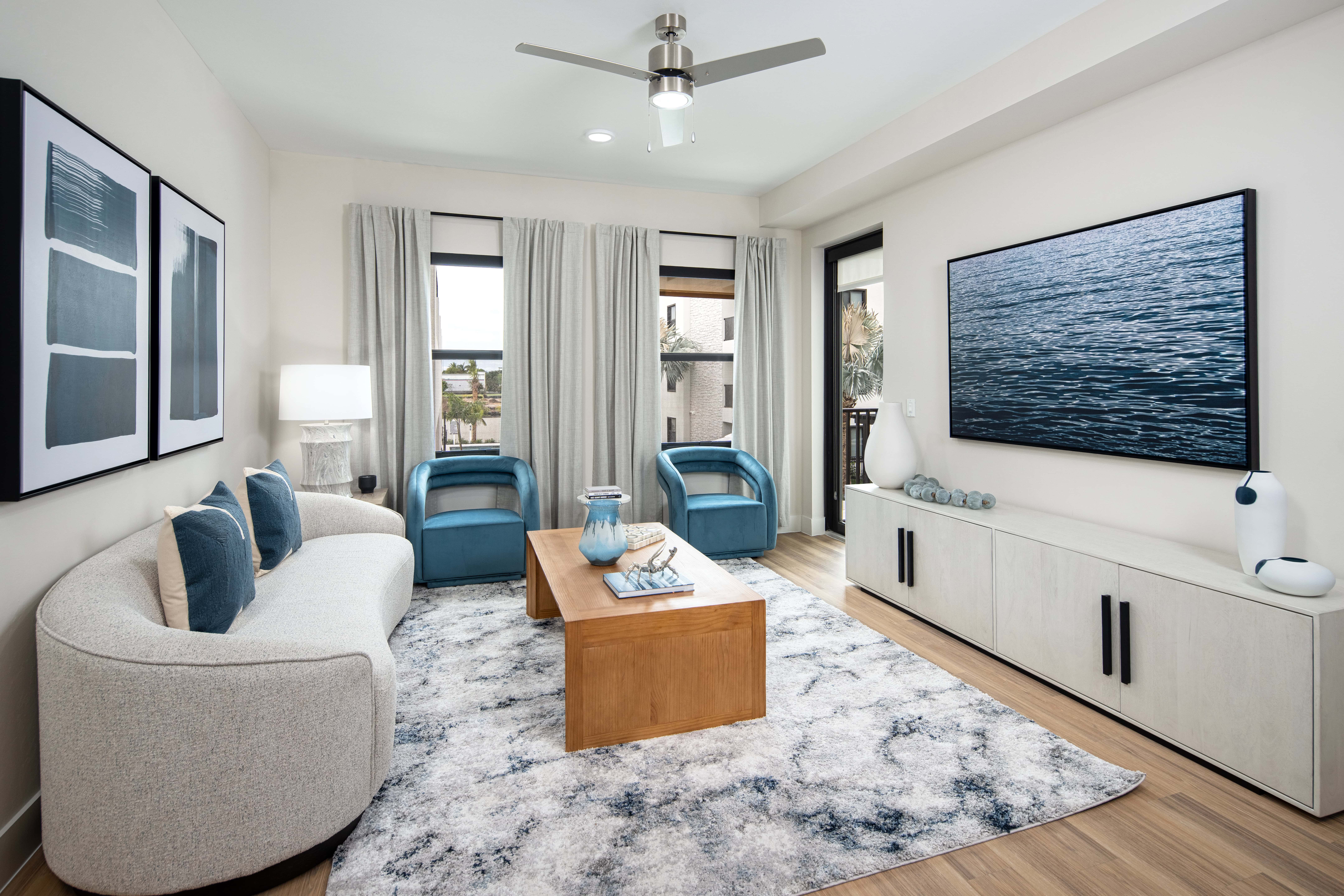 A modern living room with a minimalist design featuring a curved sofa and two blue armchairs. A wooden coffee table is placed in the center with a decorative bowl. A large flat-screen TV is mounted on the wall opposite the seating area, and the room is decorated with light-colored curtains and abstract artwork.