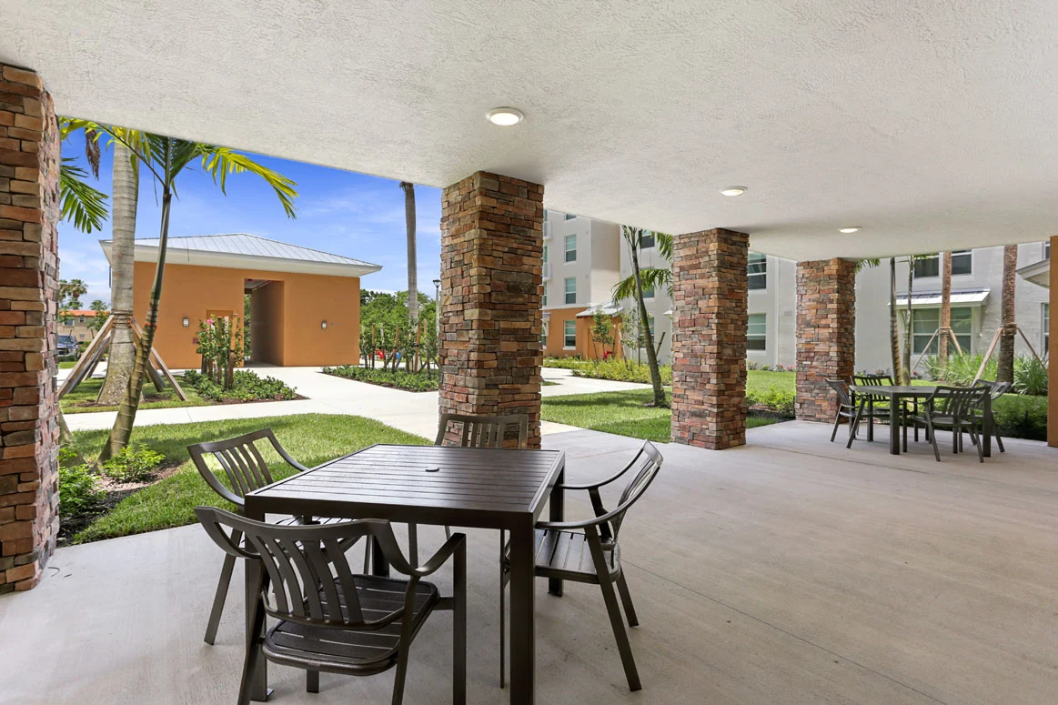 A covered outdoor seating area with tables and chairs, surrounded by palm trees and greenery.