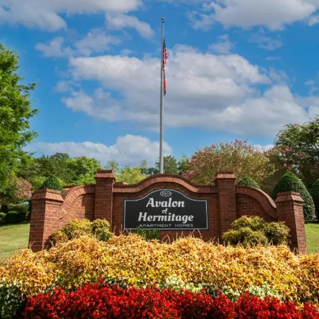 The sign for Avalon Hermitage Apartment Homes surrounded by colorful flowers and greenery on a sunny day.