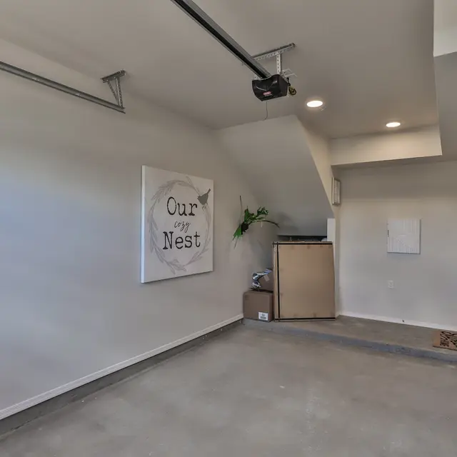 A modern, empty garage space with concrete flooring and minimal decor. A wall features a canvas art piece that reads 'Our Nest'. There is a small shelving unit and a door leading to another room.
