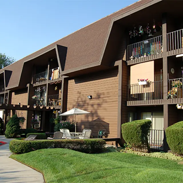 A modern apartment complex with brown wood siding and balconies adorned with plants and flowers. The area features well-maintained lawns and trees, along with a pathway and parked cars.