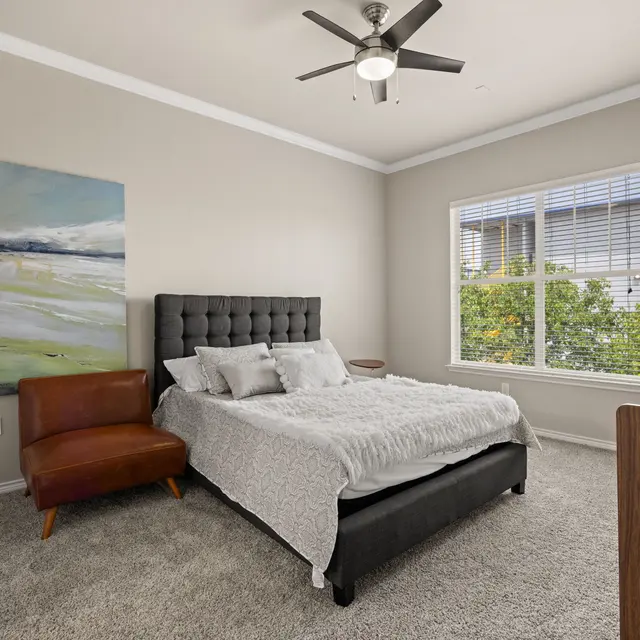 Stylish Modern Bedroom A modern bedroom with a black upholstered bed, two pillows on the bed, and a brown accent chair. A large piece of abstract art hangs on the wall, and there's a window with a view of greenery outside.