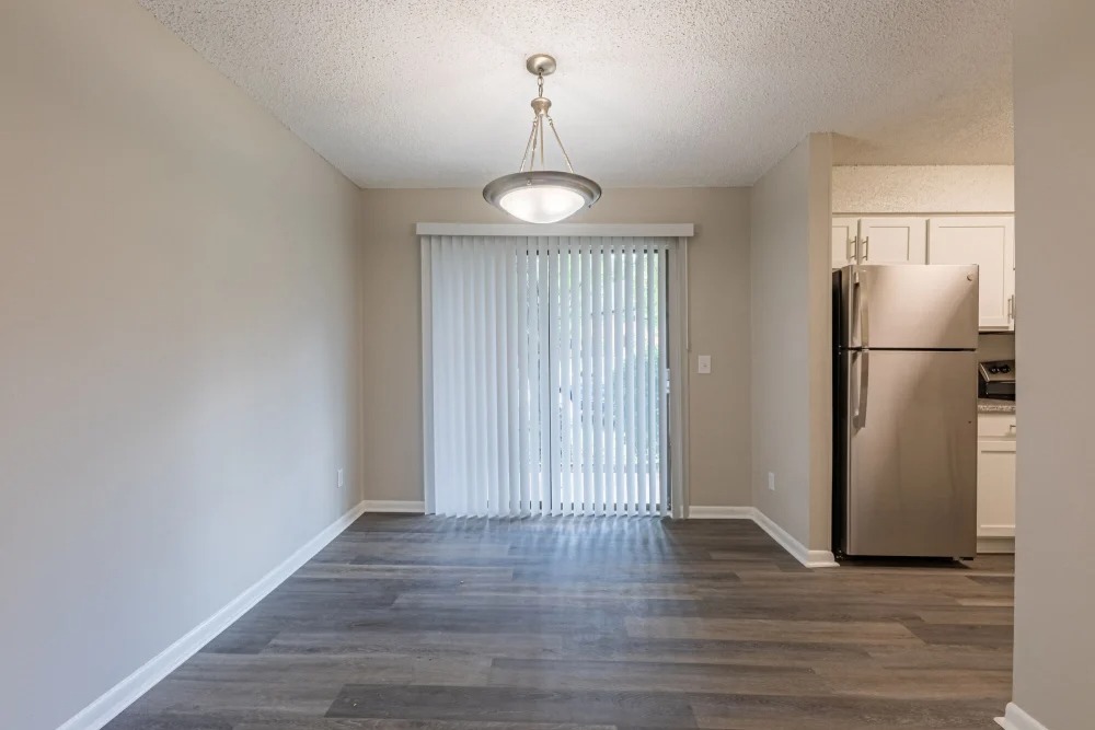 A bright dining area featuring a pendant light and sliding glass doors leading to an outdoor space. The flooring is a light wood finish, and there is a silver refrigerator visible in the adjoining kitchen.