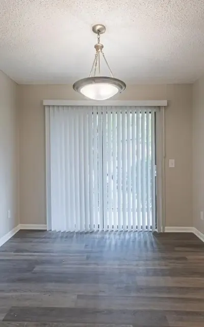 A bright dining area featuring a pendant light and sliding glass doors leading to an outdoor space. The flooring is a light wood finish, and there is a silver refrigerator visible in the adjoining kitchen.