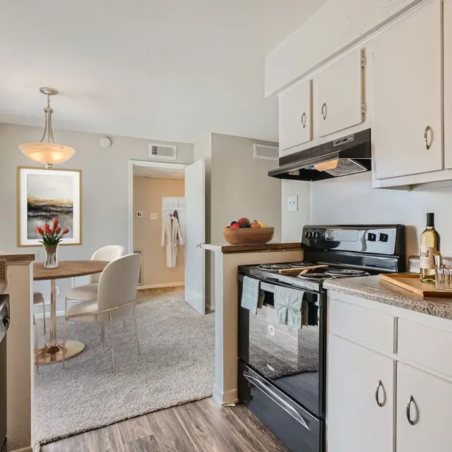 A modern kitchen featuring black appliances, white cabinetry, and a dining area with round table and chairs. Natural light is streaming through the windows.