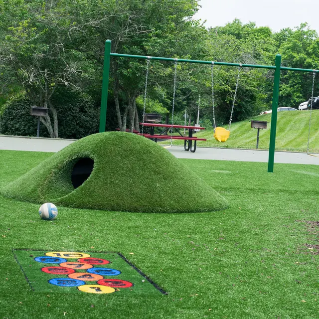 A playground with a grassy hill, a swing set, and a colorful hopscotch grid on a green turf surface.