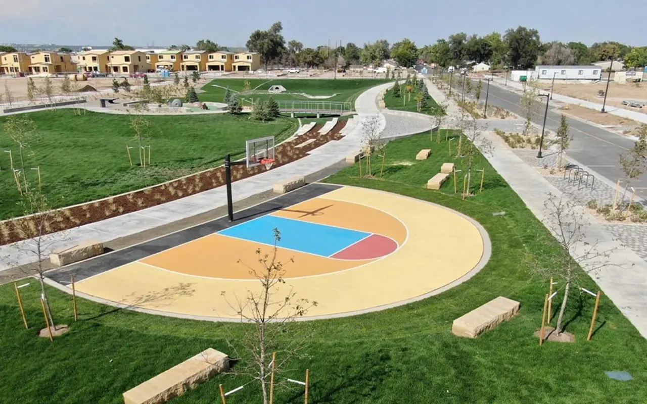 A view of a community park featuring a colorful basketball court surrounded by green grass, trees, and seating areas. In the background, there are new residential buildings and a pathway.