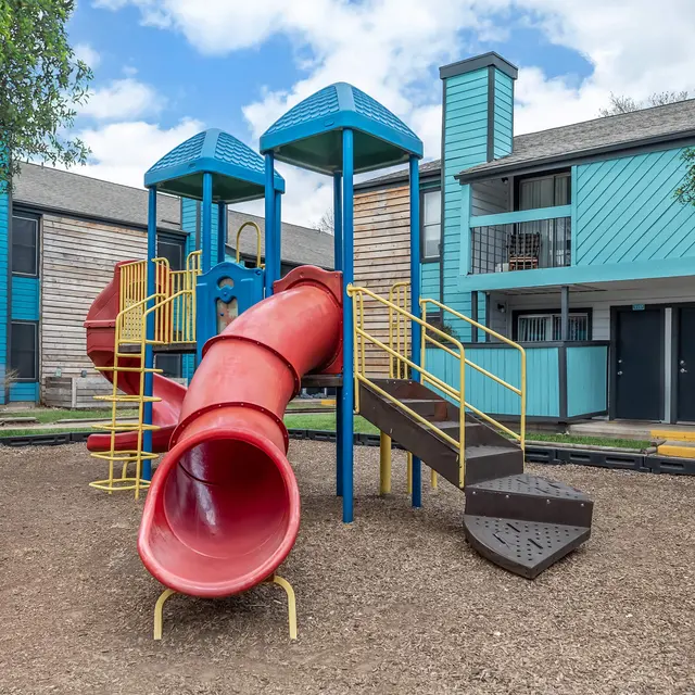 Colorful playground featuring a red slide surrounded by various play structures and apartment buildings in the background.