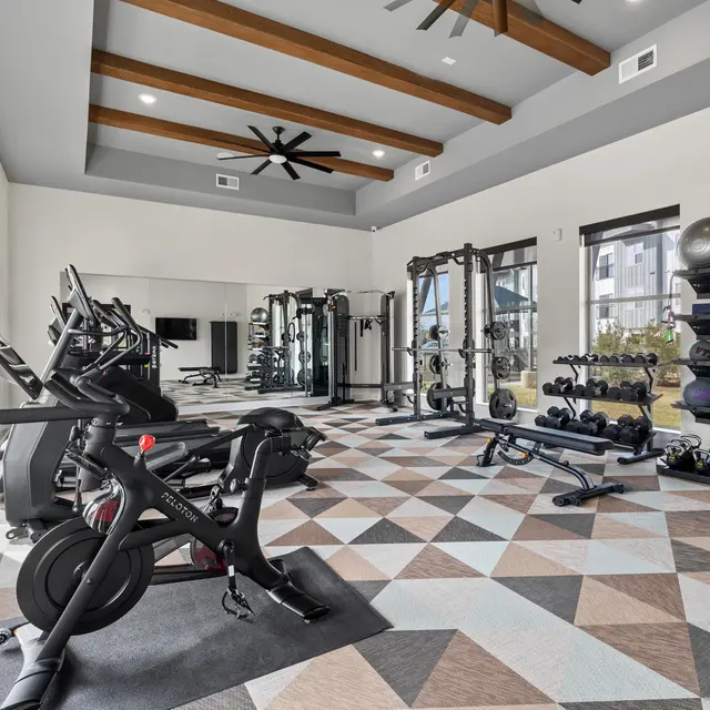A modern gym space featuring exercise equipment, including stationary bikes, weight racks, and a multi-station gym. The flooring has a geometric pattern, and large windows provide natural light.