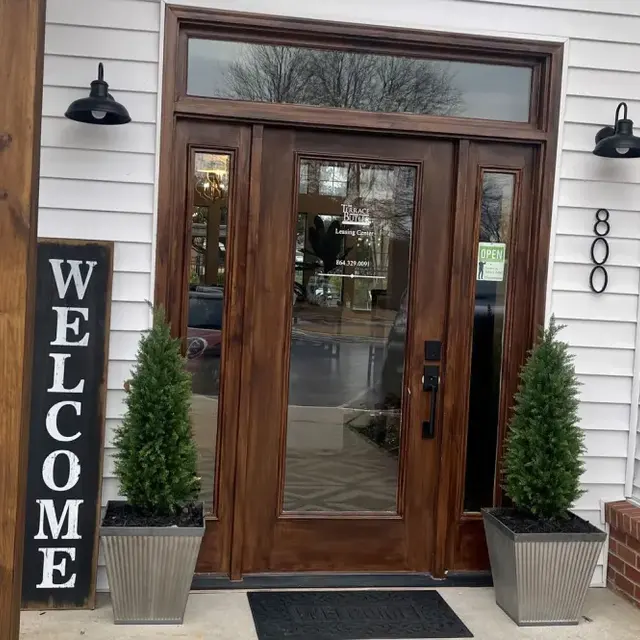 A welcoming entryway with a wooden door, flanked by two potted plants and a tall sign that reads 'WELCOME.'
