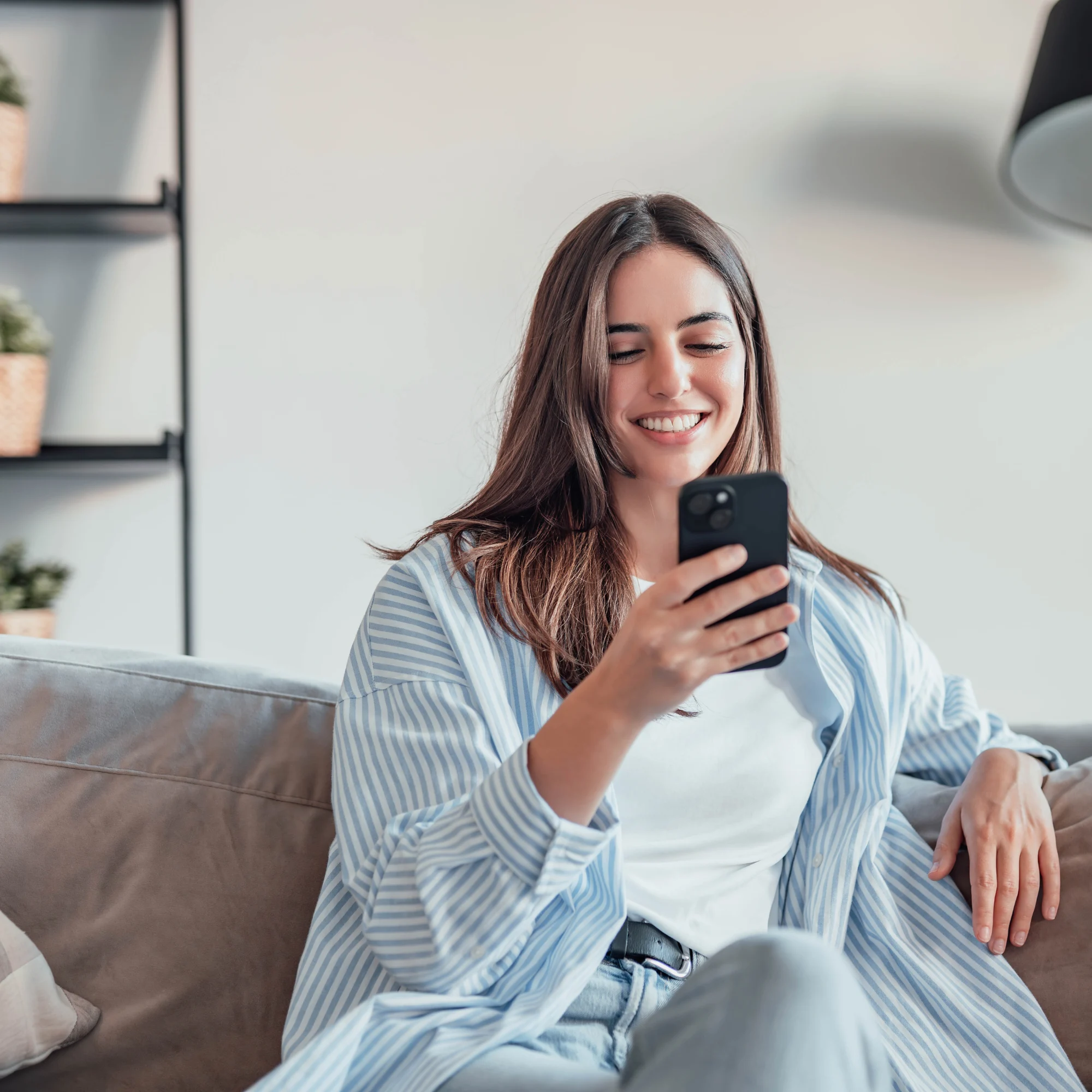 A woman sitting on a couch, smiling at her smartphone.