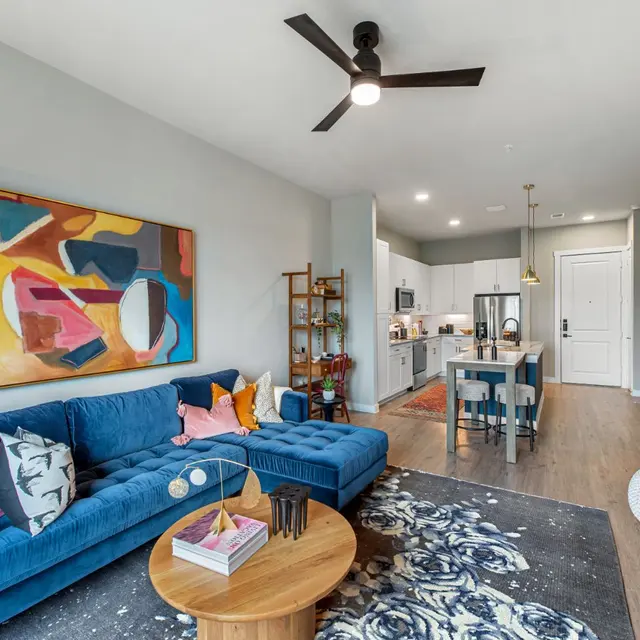 A modern living room featuring a blue sofa adorned with colorful pillows, a wooden coffee table, and an area rug. The space includes a kitchen in the background with white cabinetry and stainless appliances. There is a piece of abstract art on the wall and a decorative plant in the corner.