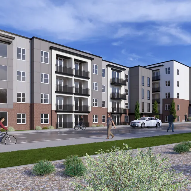 A modern apartment complex with three connected buildings, featuring balconies and large windows. People are seen walking and riding bikes along the street in front of the complex. Green grass and shrubs are in the foreground, and the sky is partly cloudy.
