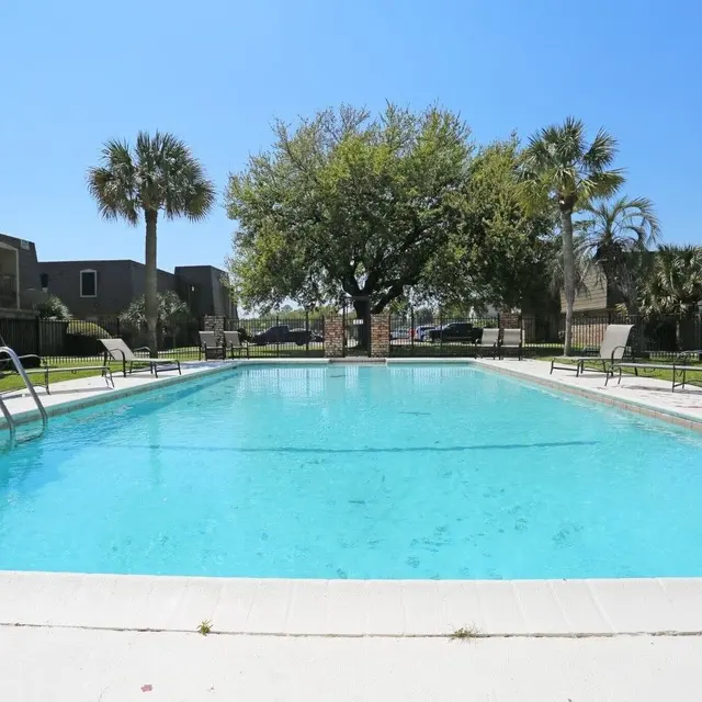 A clear swimming pool surrounded by green grass and palm trees, with lounge chairs and two buildings in the background.