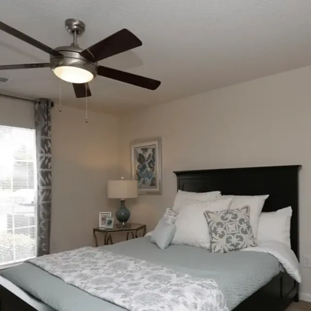 A cozy bedroom with a king-sized bed covered in a light blue blanket and decorative pillows. A ceiling fan with light hangs above. The window has sheer curtains letting in natural light, and a bedside table with a lamp and decorative items is next to the bed.