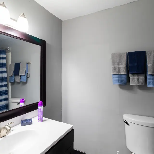 A modern bathroom with a mirror, sink, and toilet. The walls are painted gray, and there is a shower curtain with blue stripes. Two sets of towels are neatly hung on the wall, and various toiletries are placed on the countertop.