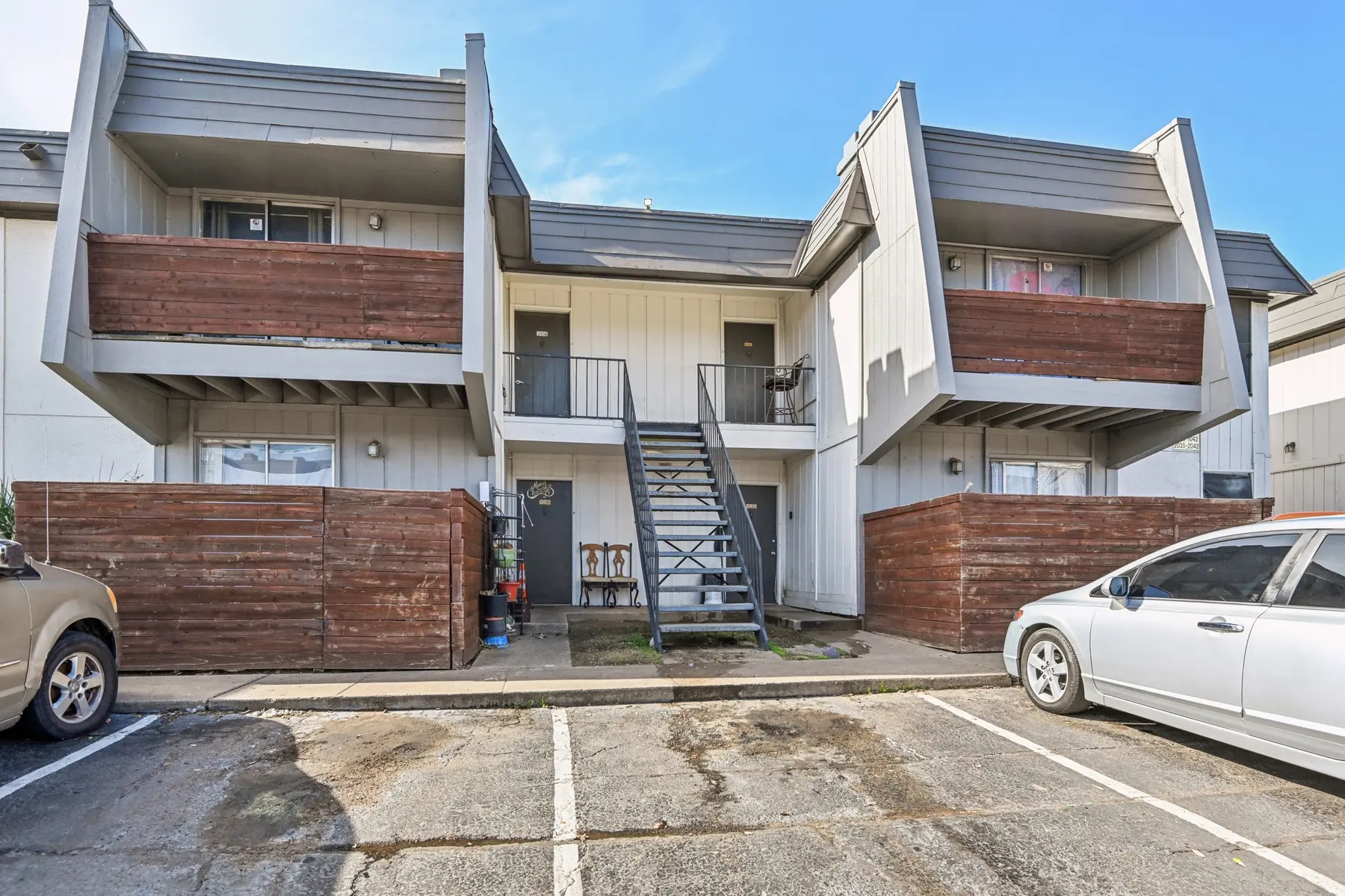 An exterior view of a two-story apartment building with wooden balconies. A set of stairs leads to the second floor, and there are two parked cars in front.