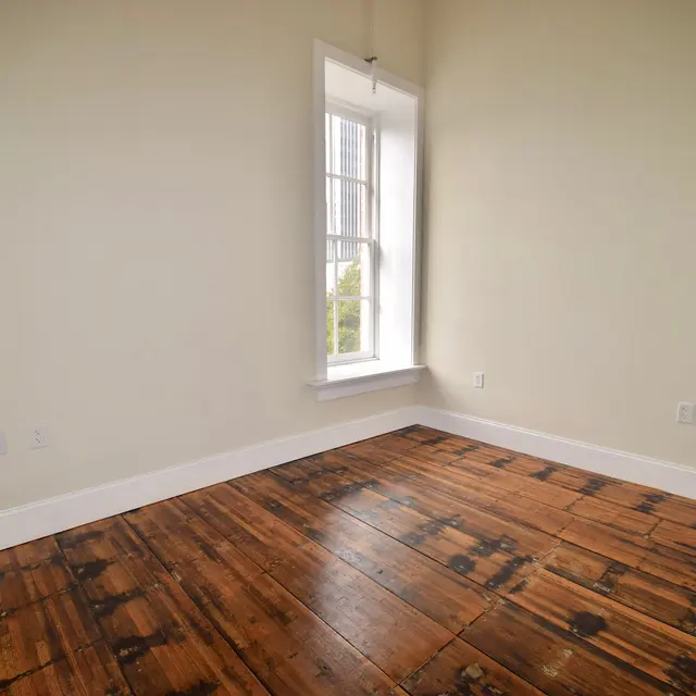 A spacious, empty room featuring a large window and wooden flooring.