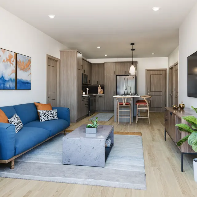 A modern living room featuring a blue sectional sofa with decorative pillows, a coffee table, and a plant. The space includes an open kitchen area with wooden cabinets and a dining table. Artwork is displayed on the wall, and there is a television mounted on another wall.