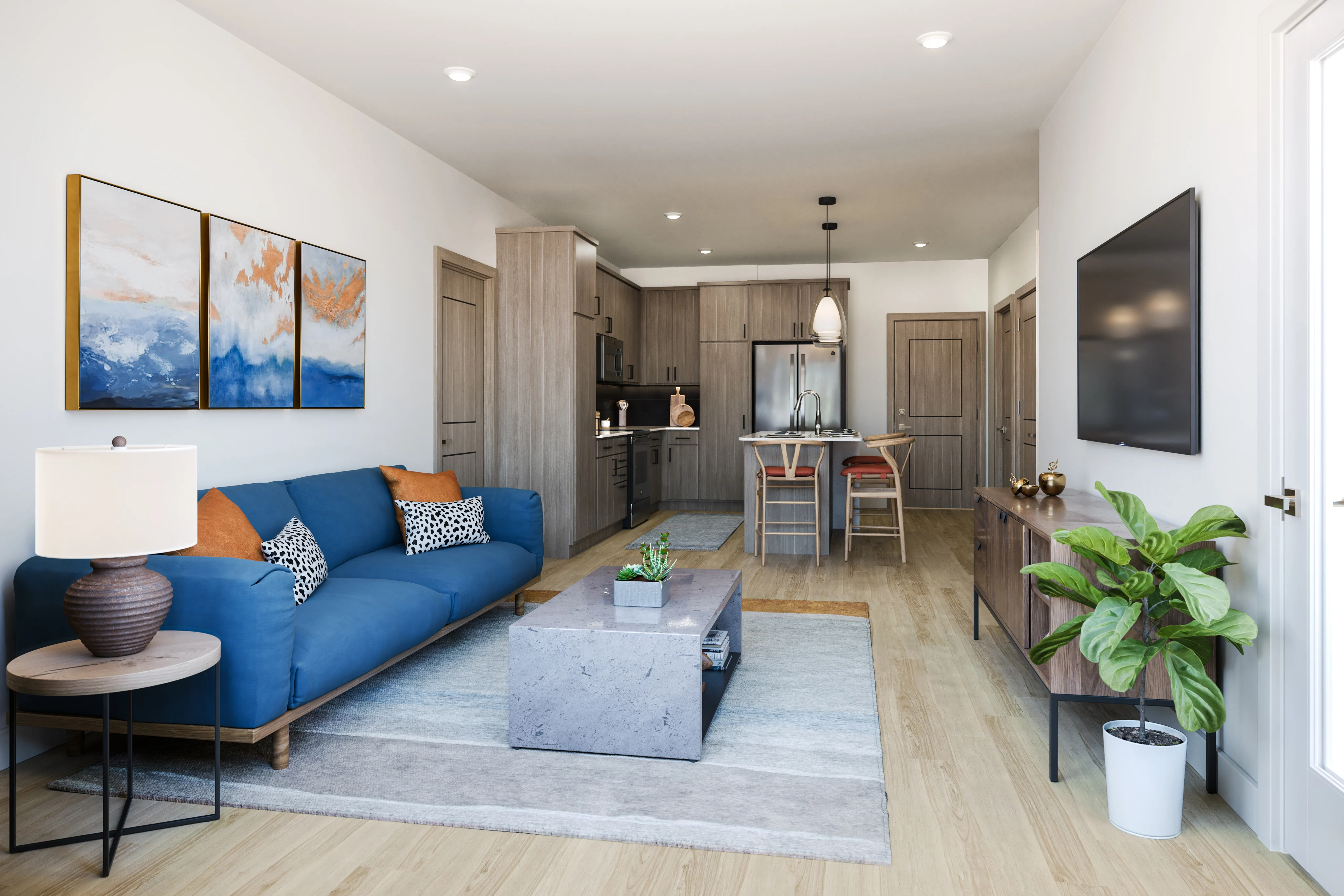 A modern living room featuring a blue sectional sofa with decorative pillows, a coffee table, and a plant. The space includes an open kitchen area with wooden cabinets and a dining table. Artwork is displayed on the wall, and there is a television mounted on another wall.