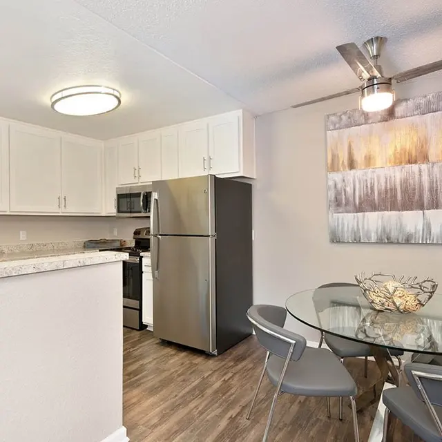 A modern kitchen and dining area featuring stainless steel appliances, white cabinetry, and a round glass dining table with gray chairs.
