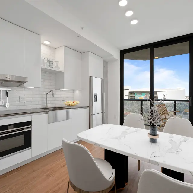 A modern kitchen with a white color palette, featuring sleek cabinetry, a marble dining table with chairs, and large windows providing a view of the outdoors.