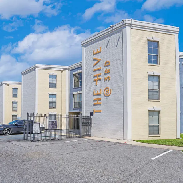 A modern apartment building named 'The Hive @ 3rd', featuring a light-colored exterior with blue accents and multiple windows. The foreground shows a parking lot with several cars and green grass.