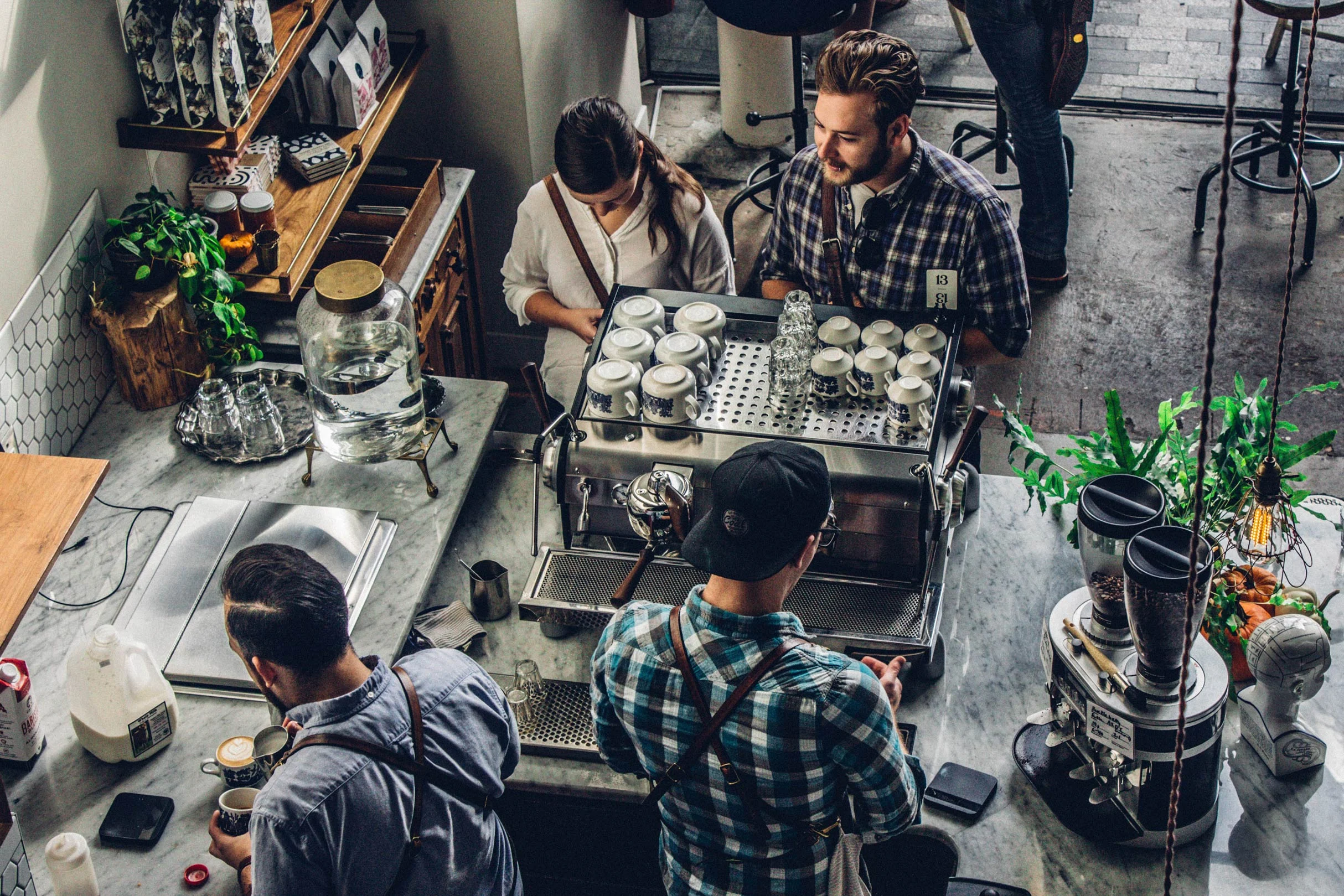 A coffee shop scene showing baristas working behind a counter. One barista prepares drinks while another takes notes on a phone. Various coffee equipment and decor can be seen in the background.