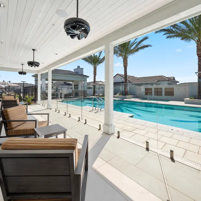 A modern patio with wooden chairs overlooks a swimming pool surrounded by palm trees and a sunny sky.