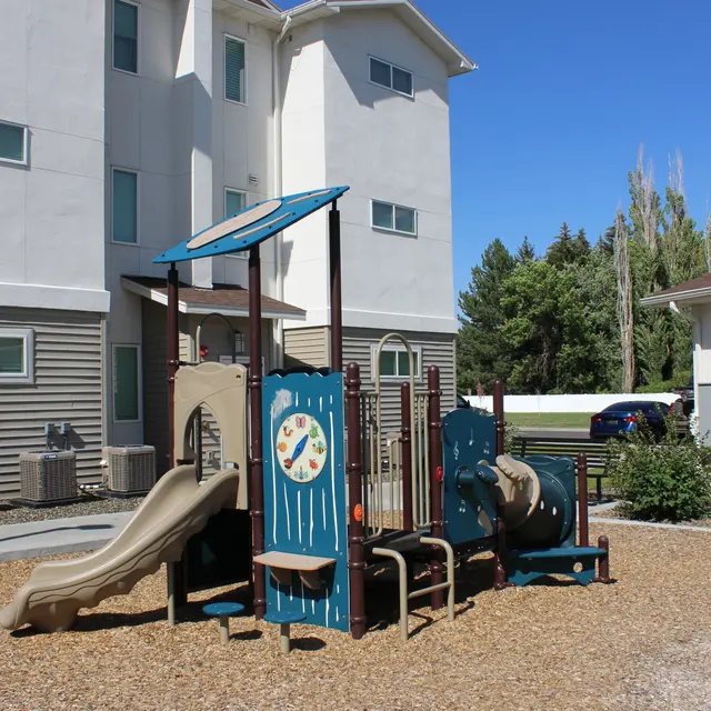 A playground with slides and climbing structures surrounded by gravel, adjacent to residential buildings.