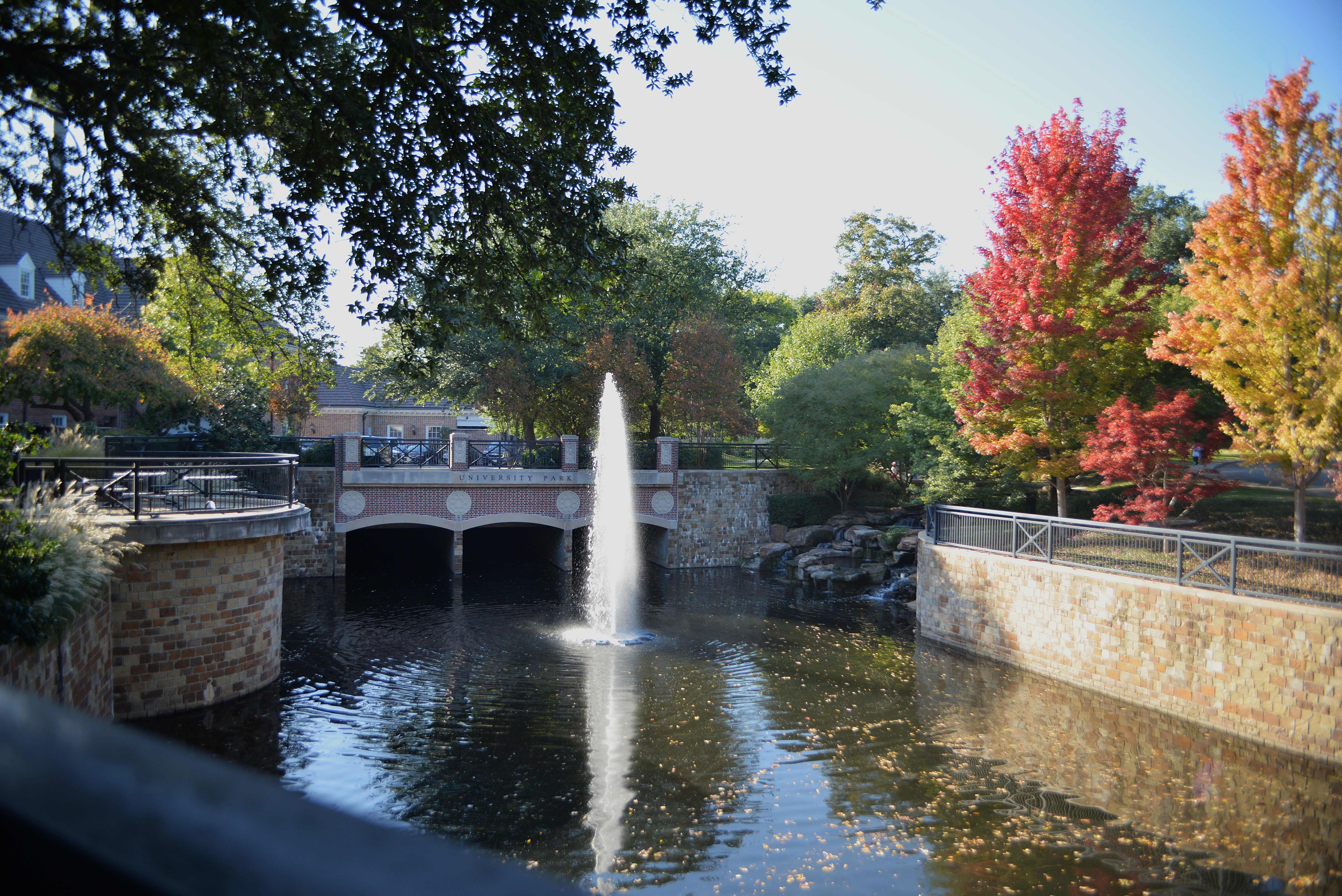 Tranquil Park with Fountain A serene park scene featuring a fountain in the center of a pond, surrounded by trees with autumn foliage in shades of red, orange, and green. A stone bridge crosses over the water, enhancing the tranquil atmosphere.