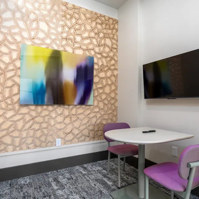 A modern small meeting room featuring a textured wall, a colorful abstract painting, and a wall-mounted television. The room includes a small table with chairs, and has a neutral colored carpet.
