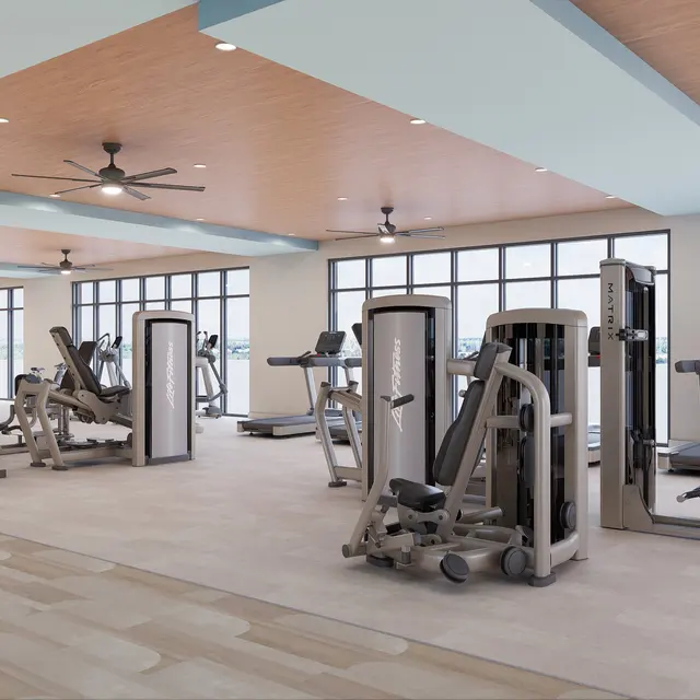A spacious, modern gym interior featuring various fitness machines and equipment. Large windows provide natural light, and the ceiling has wooden panels with ceiling fans.