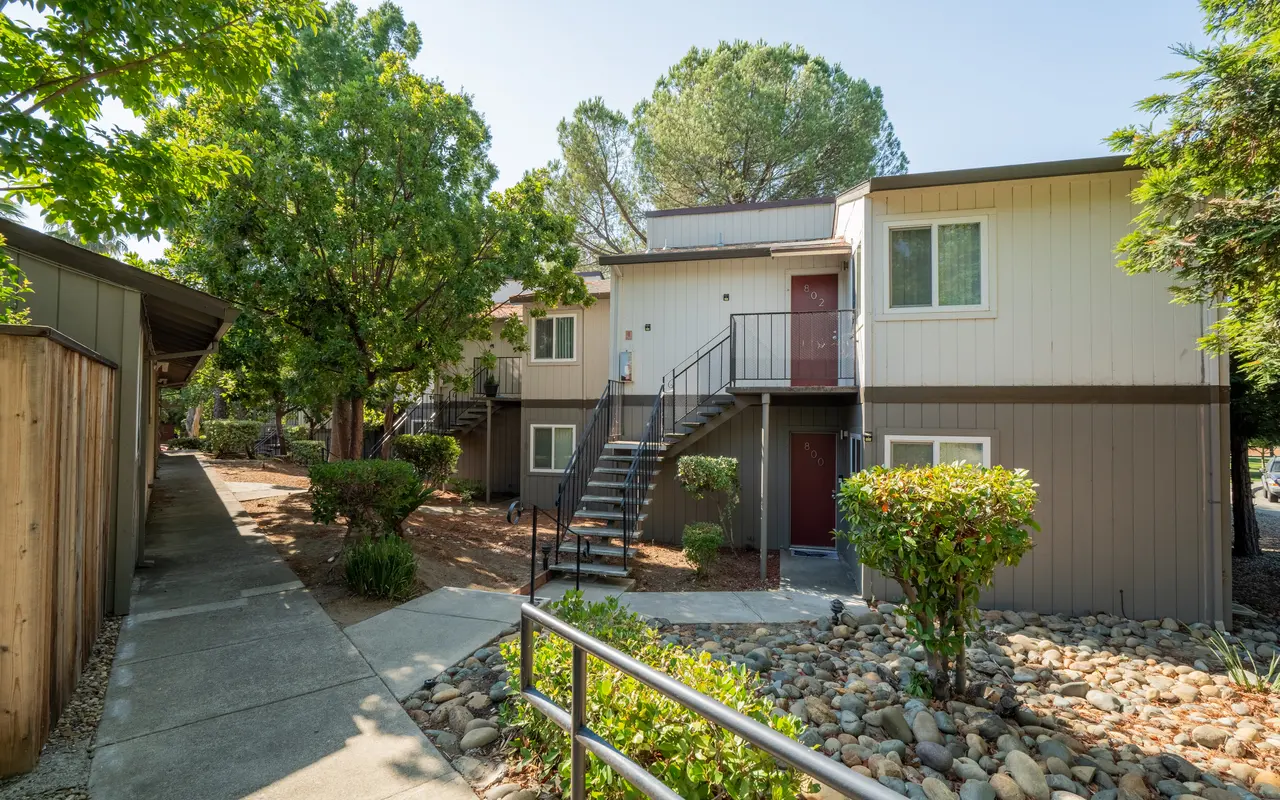 Exterior view of a multi-unit apartment complex surrounded by lush trees and a landscaped pathway.