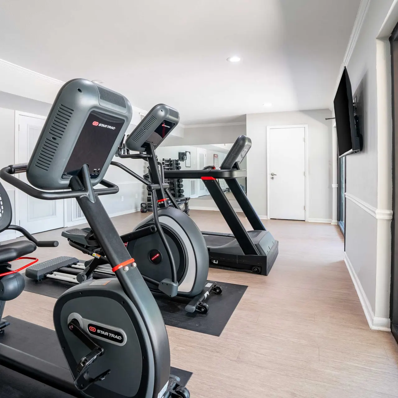 A modern fitness center with exercise equipment including a stationary bike and treadmills. The room is bright and includes large windows with views outside.