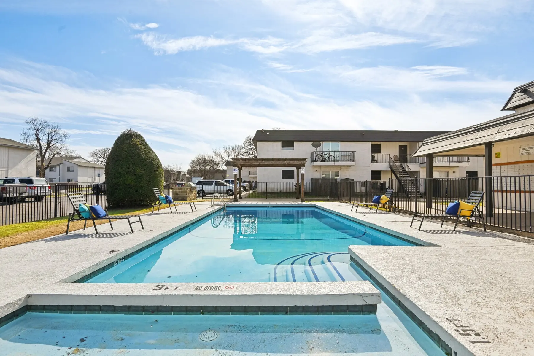 A swimming pool area with lounge chairs and a landscaped area, surrounded by a fence and buildings in the background under a partly cloudy sky.