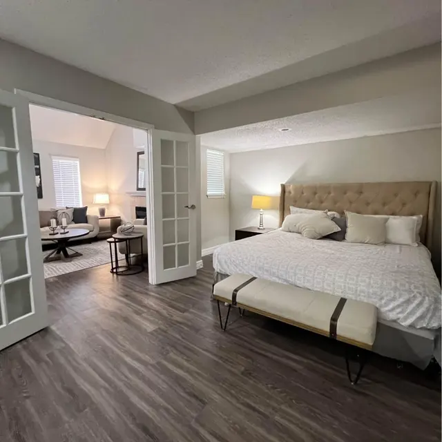 Cozy Modern Bedroom A cozy bedroom with a king-sized bed, light-colored walls, and hardwood flooring. Through a set of French doors, a sitting area with a round table and chairs is visible. Soft lighting creates a warm ambiance.