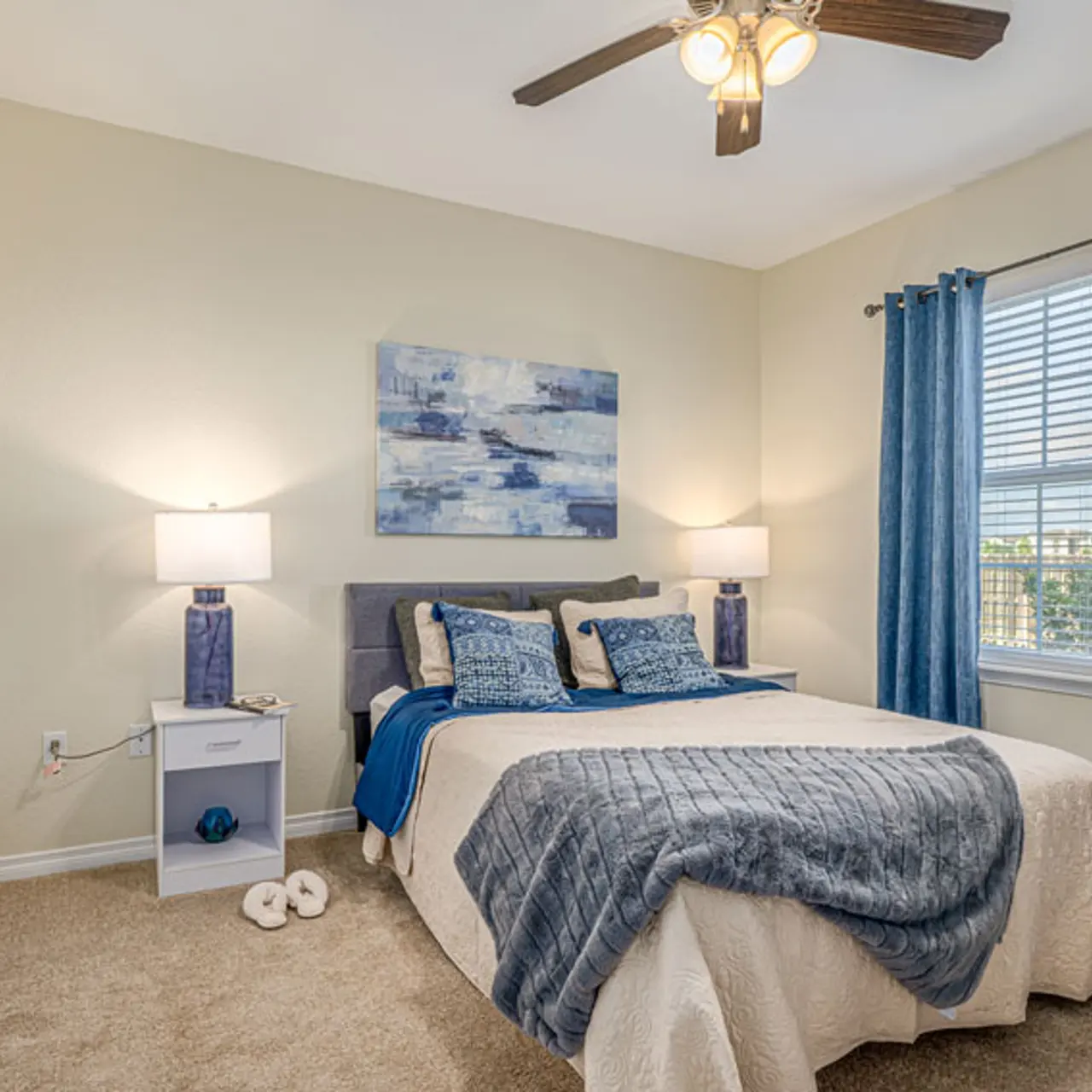 A cozy bedroom featuring a queen-sized bed with blue and gray bedding, two lamps on nightstands, and large windows with blue curtains.