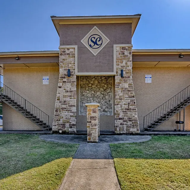 Front view of a two-story apartment building with exterior staircases on each side. The building features a combination of stone and beige siding, with a decorative sign containing the letters 'SC' at the top.
