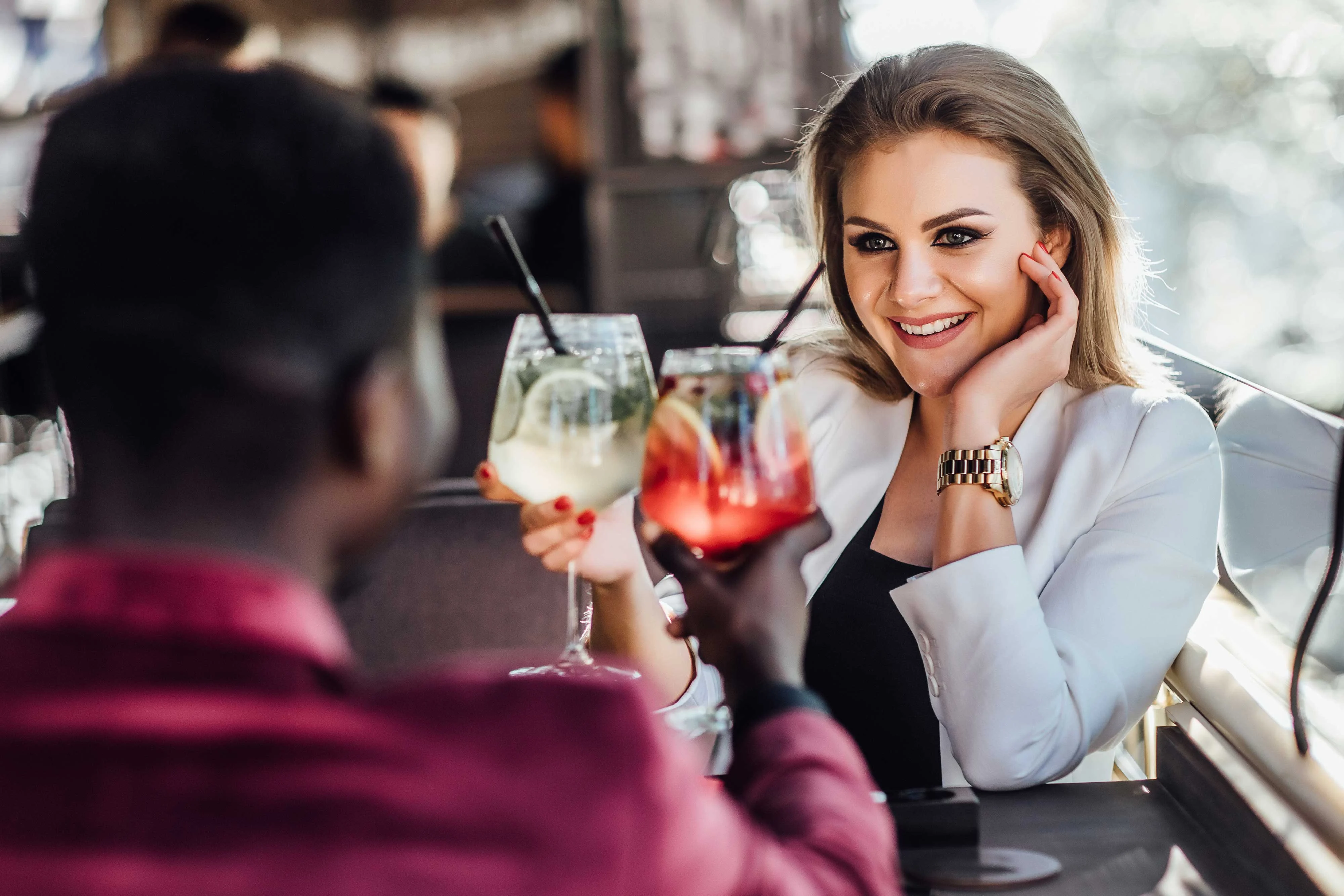 A couple enjoying a drink at a cafe. The woman is smiling, holding a colorful cocktail, while the man is facing away.
